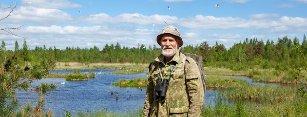 Naklejka premium A man with a beard in camouflage clothes, with binoculars, an explorer, a tourist. Stands against the background of a forest lake, forest and blue sky with clouds.