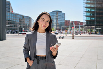 Young smiling elegant Asian busy business woman leader wearing suit standing in big city using cell...