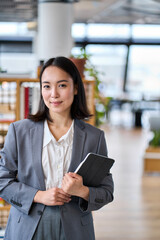 Smiling young Asian business woman wearing suit holding digital tablet standing in office....