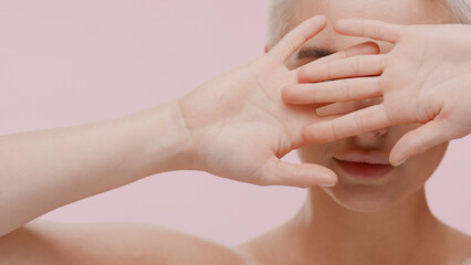 Naklejka premium Beautiful young slim white-skinned woman with fair hair covers her face with her hands on pale pink background | Face skin protection concept