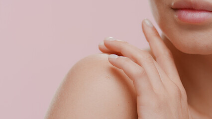 Extreme close-up shot of young slim good-looking white-skinned female model strokes her naked shoulder on pale pink background | Soft skin concept