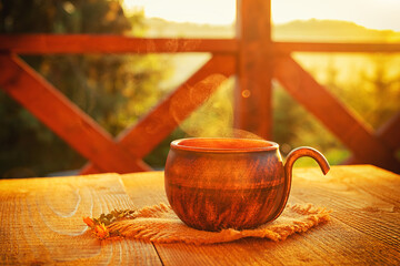 handmade brown ceramic cup with herbal tee on wooden table. Goldish morning sunlight comes  through steam over the cup.