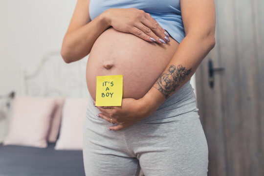 Gender reveal concept. It's a boy. Yellow sticker placed on a caucasian pregnant person's belly. Tattoo on arm, long nails, biker shorts. Middle body bart photo. High quality photo
