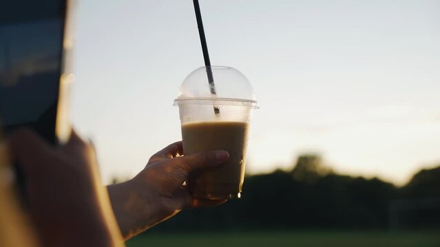 The girl holds a cup of coffee in her hand and takes pictures of it. She takes pictures against the sunset