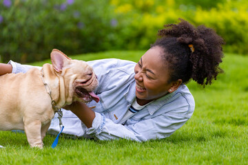 African American woman is playing with her french bulldog puppy while lying down in the grass lawn after having morning exercise in the park