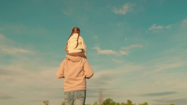 Child sits on fathers neck, Dad walks with his daughter on his shoulders against blue sky, paternity. Happy family. Father baby girl are walking together in park.Carefree family, dad, daughter travel