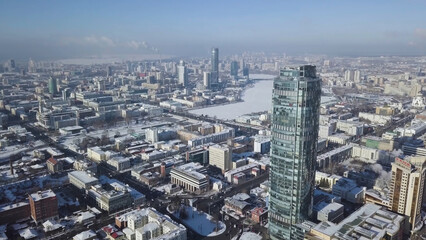 Fototapeta na wymiar Top view of the amazing glass tower or the business center in the background of a winter city. Aerial view of skyscraper is in the middle of the city in winter, blue sky sky and snowy roofs of