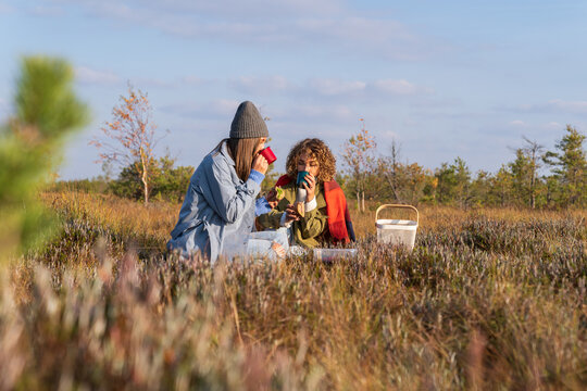 Two trendy girls having cozy autumn picnic in countryside with hot tea, sandwiches enjoying sunny days. Tea break. Happy laughing female friends or sisters travel to woods on weekend together