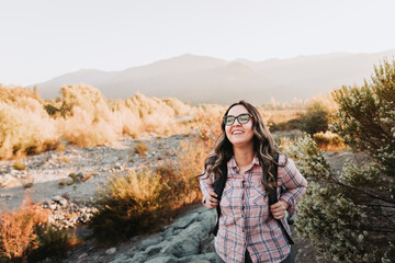 Smiling happy latin young woman with glasses and a backpack on, hiking in a beautiful landscape.
