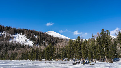 Two people are standing next to a snowmobile in a snowy valley. Traces of runners in the snow. Coniferous forest grows on the hillside. The top of the mountain against a clear blue sky. Altai