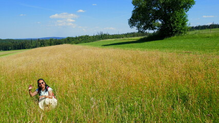 junge Frau mit Sonnenhut posiert sitzend im hohen Gras mit Baum und grüner Wiese im Schwarzwald...