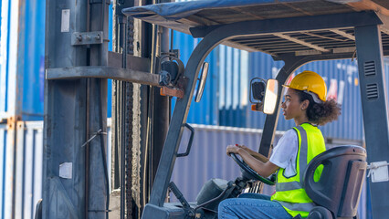 Naklejka premium Young African American lady operates a forklift tractor to move containers around the shipyard. Young female worker with yellow helmet, and safety suit is driving a truck
