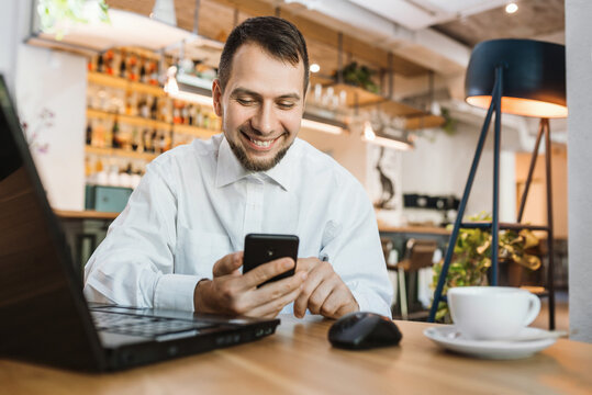 Smiling modern young bearded man using gadgets in cafe. Businessman working while taking a break, no time to waste
