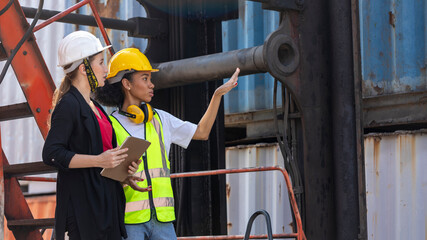 Two female logistic workers from different cultures or ethnicities have conversation next to shipping blue containers. A female foreman discusses with cargo female worker for stocking