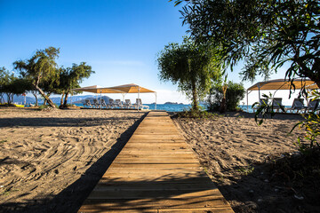 A wooden path in the sand goes to the beach with umbrella, sun-beds and chairs