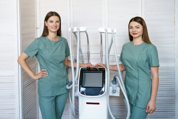 Two women specialists in hardware cosmetology stand in massage room at laser therapy apparatus....