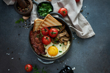 Fried eggs, bacon, tomato and toasted bread in a black ceramic pan at domestic kitchen