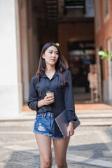 Beautiful Asian woman holding a cup of coffee with a laptop. and standing in front of the office building to go to work in the morning