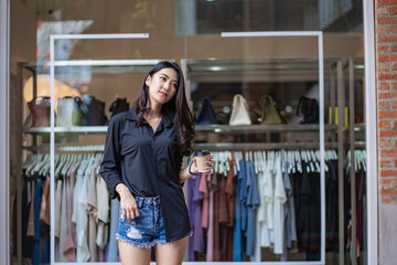 A beautiful Asian woman holding a cup of coffee in a black shirt is smiling happily and standing in front of the holiday concept building.