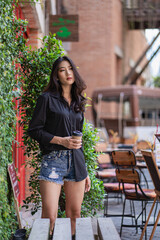 A beautiful Asian woman holding a cup of coffee in a black shirt is smiling happily and standing in front of the holiday concept building.