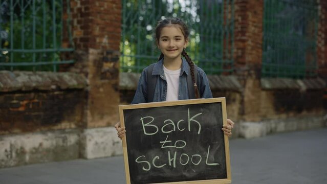 Portrait of little girl standing turn around smilling and holding schoolboard with text back to school on the street. Female school student looking at the camera. Education concept
