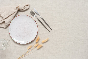 Autumn table setting. A white plate, cutlery, a beige linen napkin and dry grass lagurus on a beige linen tablecloth. Top view, flat lay. 