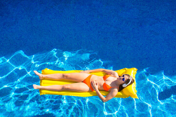 Aerial view of a young woman in her 20s wearing an orange bikini, lying on a yellow inflatable mat inside the pool. She is listening to music with white headphones. Blue background. Lifestyle - Powered by Adobe