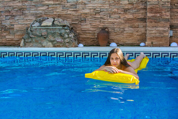 Young woman in her 20s smiling. Blonde girl in the pool wearing an orange bikini lying on a yellow...