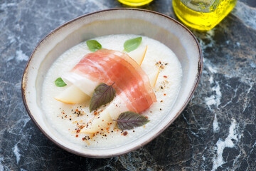 Melon cream-soup topped with jamon and fresh basil leaves, horizontal shot on a black marble background, middle close-up