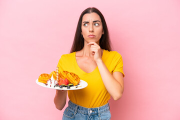 Young caucasian woman holding waffles isolated on pink background having doubts and thinking