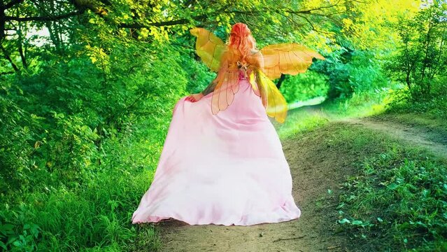 Portrait fantasy woman faerie with butterfly wings walks on path back rear view. Girl fairy tale angel, Summer nature green yellow leaves forest. Pink dress long hen flutter fly in wind slow motion