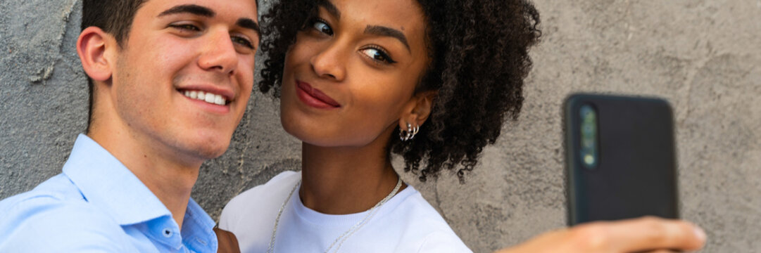 Mixed race young couple taking a selfie with smartphone – happy multiethnic couple taking photo – loving couple selfie in the city - banner size for header design