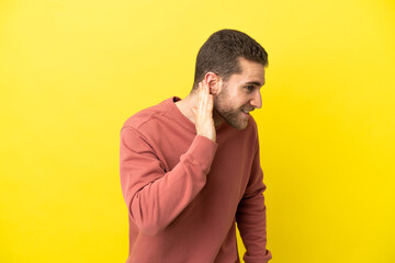 Handsome blonde man over isolated yellow background listening to something by putting hand on the ear