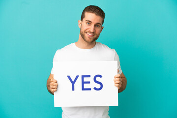 Handsome blonde man over isolated blue background holding a placard with text YES with happy expression