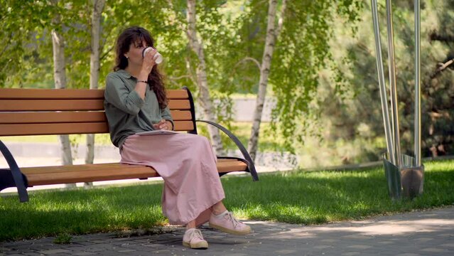 A girl in a pink skirt in the park on a bench drinks coffee.