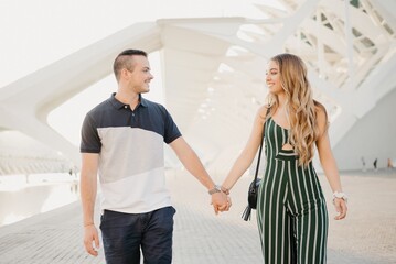 A couple of tourists is strolling in the modern urban space on a date at the sunset in Valencia. A guy is holding hands with his girlfriend in the evening in Spain.