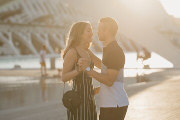 A couple of tourists is dancing in the modern urban space on a date at the sunset in Valencia. A smiling girl is holding hands with her romantic boyfriend in the evening in Spain.