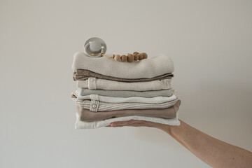 Person's hand holding cozy hygge pastel clothes and accessories for newborn baby. Basic bodysuit,...
