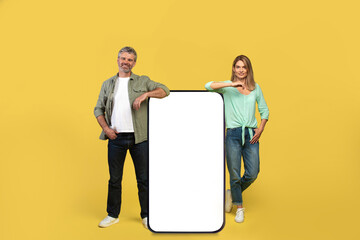 Happy middle aged couple leaning on giant smartphone with blank screen mockup, advertising your service or product