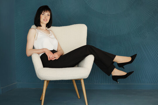 full body side profile view of smiling young brunette woman lifting legs arms up sitting and relaxing on chair on blue wall background. Female in black pants, white blouse and classic high heels shoes