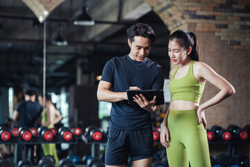 Asian man and woman exercise training in fitness gym. Young male trainer using tablet for schedule exercise training to fitness female. Workout training in fitness gym. - Powered by Adobe