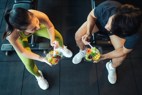 Top view Asian man and woman healthy eating salad after exercise at fitness gym. Asian couple eating salad for health together. Selective focus on salad bowl on hand.