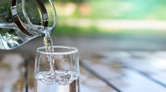 Water from jug pouring into glass on wooden table outdoors.Drink water pouring in to glass over sunlight and natural green background.Photo select focus with copy space.