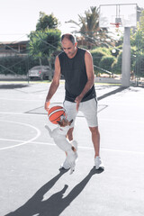 Fototapeta premium Portrait of adult senior man playing sport basketball ball with a dog jack russel terrier on playground outdoor. A pet jumping to catch the ball.