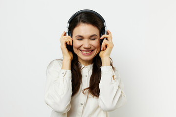 Fototapeta premium funny woman in light shirt stands listening to music in her headphones, closing her eyes with pleasure and holding headphones with her hands. Horizontal photo on a light background with an empty space