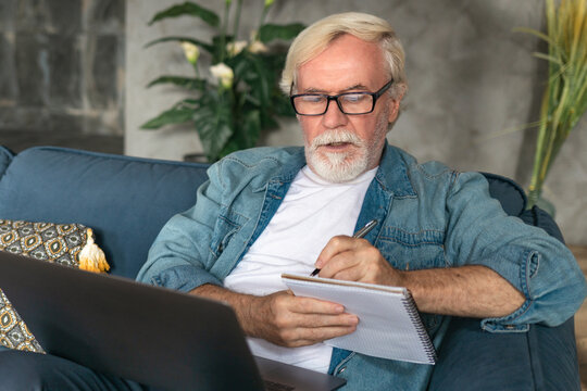 Modern Senior Businessman With Gray Hair Writing In A Notebook, Working From Home. Focused Elderly Man Writing Notes In Notebook Watching Online Webinar Using Laptop Computer