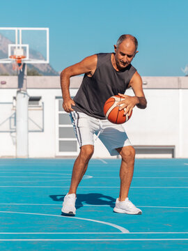 Adult senior man playing a sport ball on blue basketball court at summer sunny day.