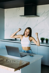 Young excited woman showing triumph gesture while talking on smartphone near laptop in kitchen