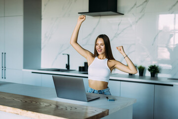 Fototapeta na wymiar Young excited woman showing triumph gesture while talking on smartphone near laptop in kitchen