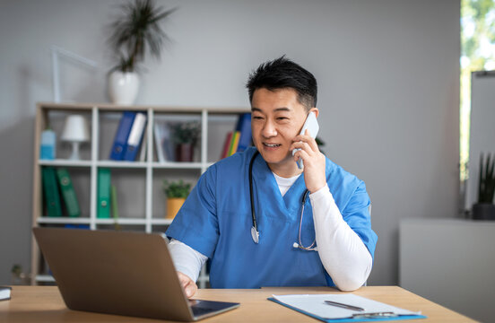 Smiling millennial asian man doctor in uniform call by phone, types on computer in office clinic interior